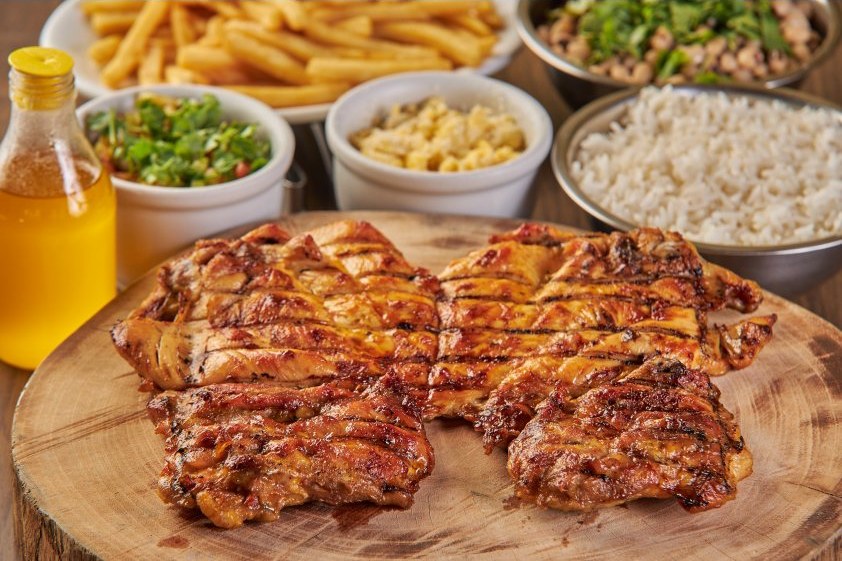 Frango grelhado na brasa servido numa tábua de madeira, com fritas, arroz, feijão verde, macaxeira e vinagrete.